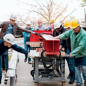 students and professors in hard hats moving a large red I-beam